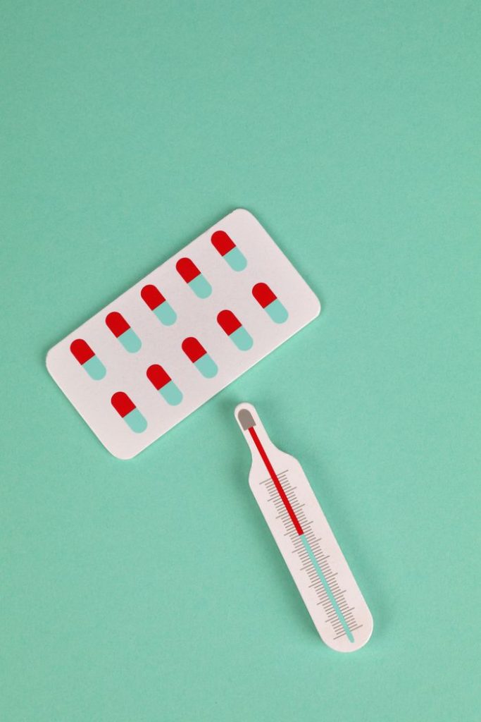 A flat lay image featuring a blister pack of pills and a thermometer on a green background, highlighting healthcare concepts.