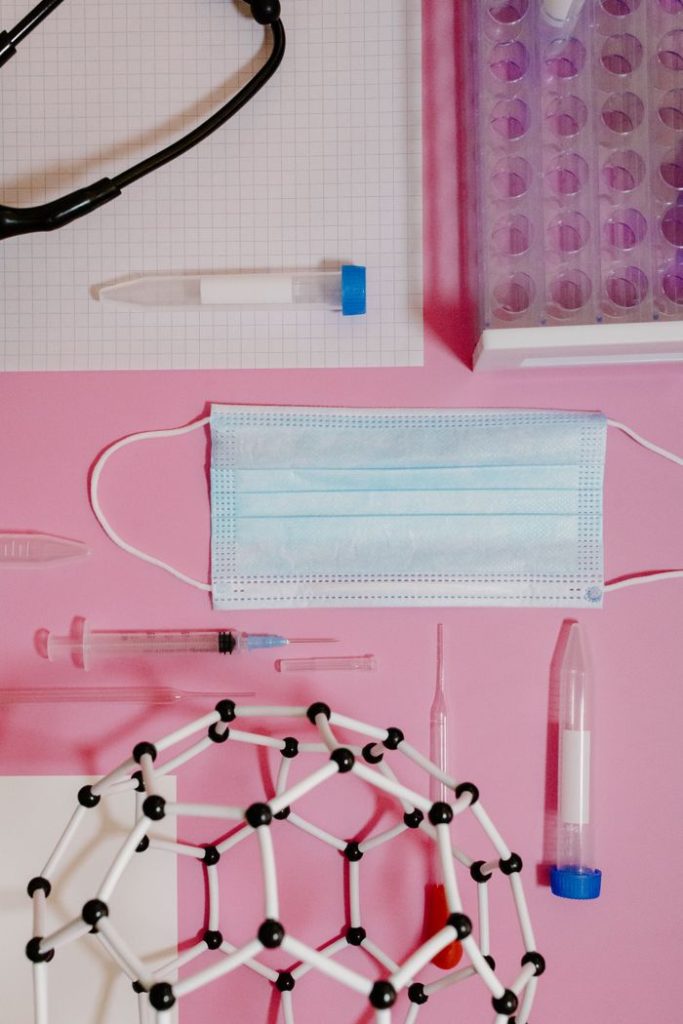 Flat lay of medical and scientific tools including mask, syringes, and molecular model on pink surface.